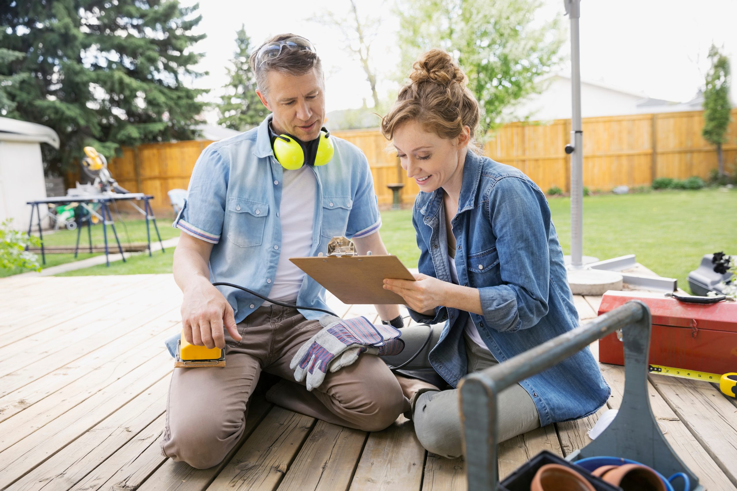 Couple planning a deck sanding project on a timber patio in East Barnet, man holding a palm sander while reviewing a checklist.