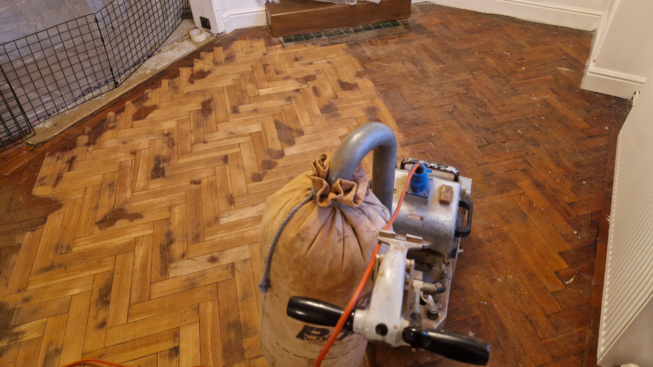 Belt sander on a worn oak herringbone parquet wood floor mid-restoration — Herringbone Parquet Sanding in East Barnet with unsanded edges visible.